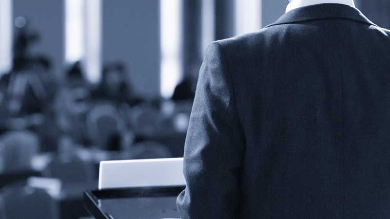 Faceless dude in a suit coat pictured from the back standing at a lectern addressing a blurry crowd of people.