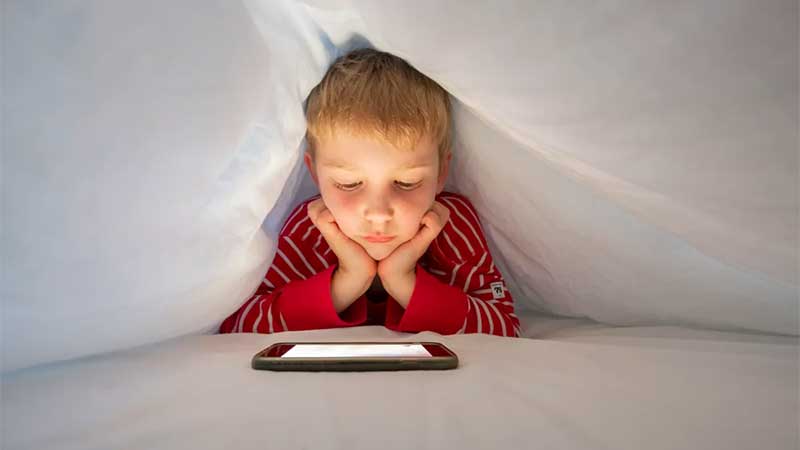 Young boy reading on a tablet under the covers.